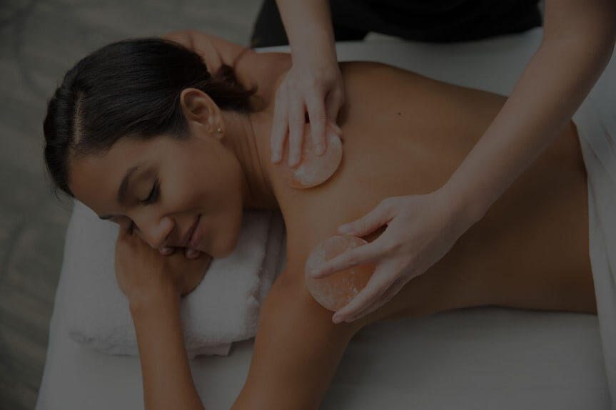 Person receiving a back massage from a therapist using soft pink salt stones.